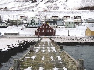 A snow-covered wooden pier leads to a village with colorful houses on a snowy hillside.
