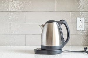 A stainless steel electric kettle on a white countertop with a tiled backsplash and an electrical outlet.
