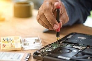 A person's hand uses a small screwdriver to repair the internal circuit board of an open electronic device, with screws visible nearby.