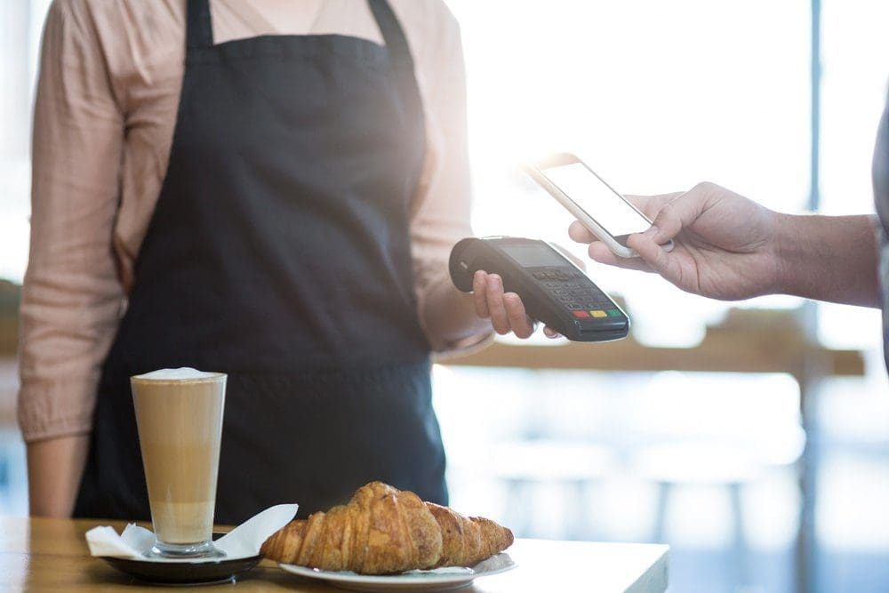 A person's hand holding a smartphone near a payment terminal for a contactless payment, with a latte and two croissants on a table.