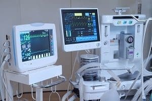 Two medical monitors displaying vital signs and waveforms next to a large white anesthesia or ventilator machine in a clinical setting.