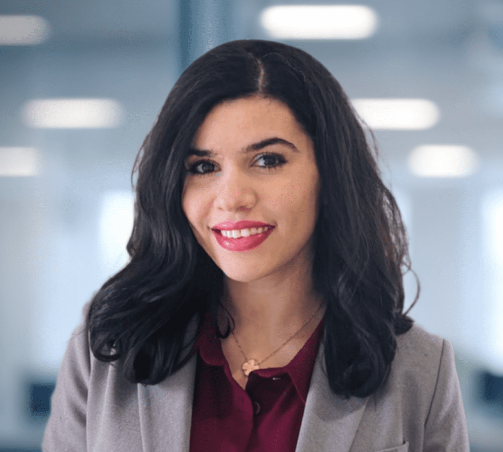 A smiling woman with dark hair and a grey blazer in an office.