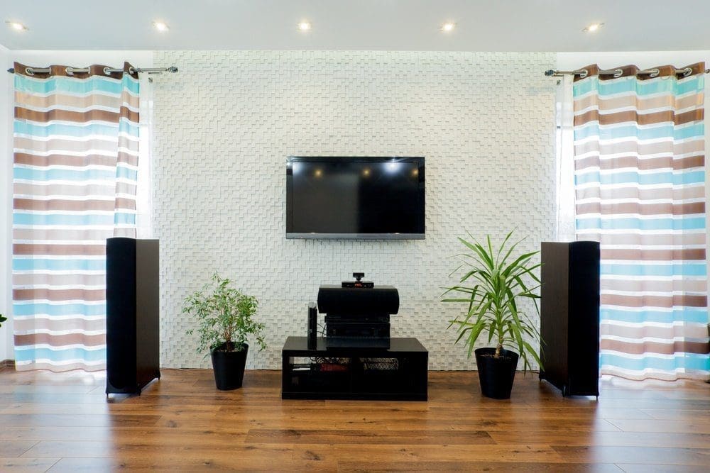 A living room with a wall-mounted TV, black media console, two tall speakers, and potted plants on hardwood floor. Striped curtains frame the setup.