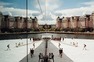 A modern waterfront plaza with a large reflective glass panel mirroring buildings, people, and a cloudy sky.