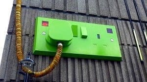 A large, bright green electrical wall socket with a plug and corrugated cable attached to a gray building facade.