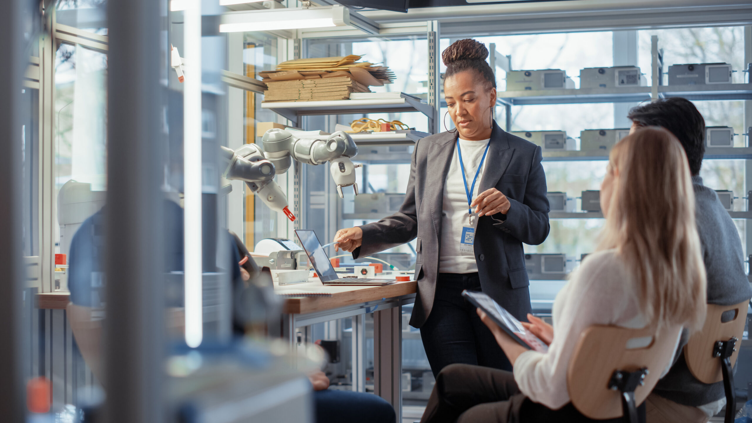 An African American woman in a blazer points at a laptop on a workbench next to a robotic arm, while two colleagues observe in a high-tech lab.