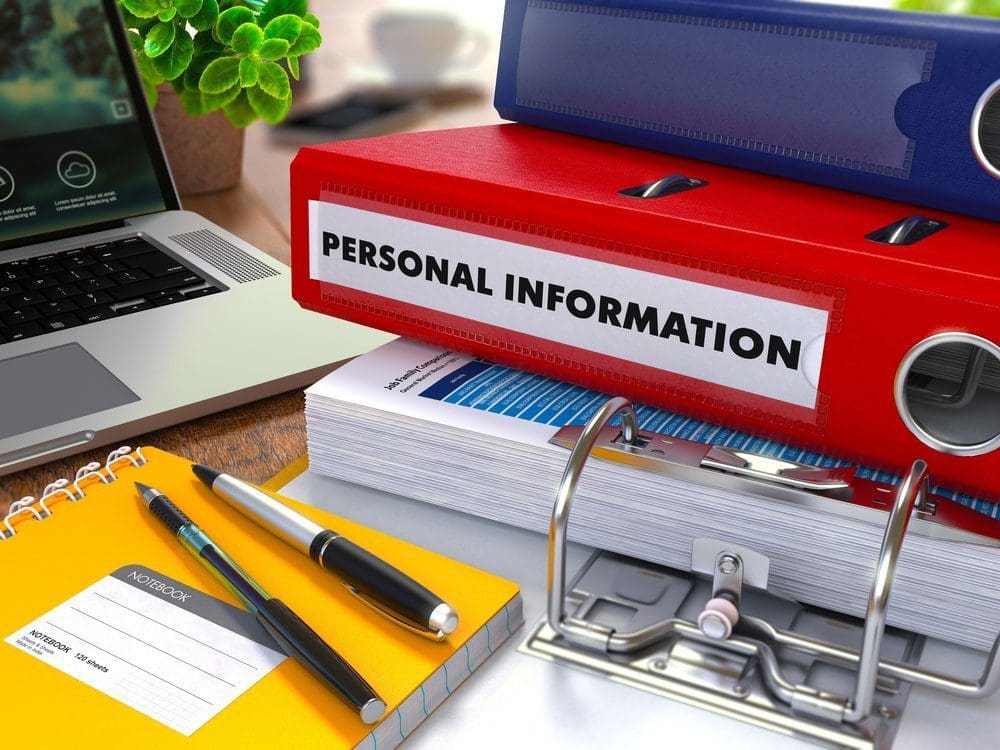 A red binder labeled 'PERSONAL INFORMATION' on a desk with a laptop, yellow notebook, pens, and other binders.