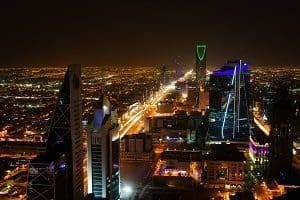 Nighttime aerial view of a modern city skyline with illuminated skyscrapers and a brightly lit highway stretching into the distance.