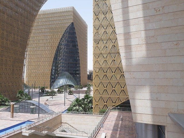 A complex of modern buildings with golden, diamond-patterned mesh facades and large glass sections, surrounding a plaza with palm trees and a water feature.