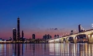 Night view of a city skyline with a brightly lit bridge over water, reflecting city and bridge lights.