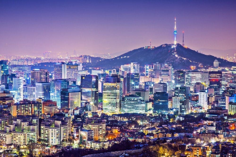 Nighttime aerial view of Seoul, South Korea, showing a vast array of illuminated skyscrapers and buildings, with Namsan Mountain and N Seoul Tower in the background against a purple sky.