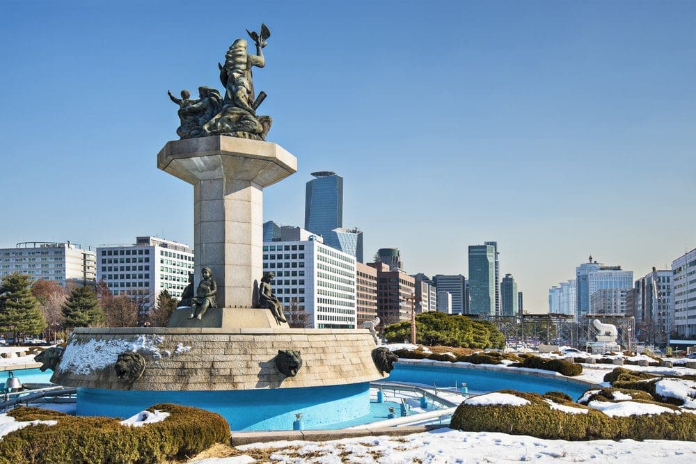 A large bronze statue on a stone pedestal with a blue fountain in front, surrounded by snow and green bushes, with a city skyline in the background under a clear sky.