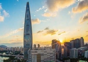 A tall, pointed skyscraper stands over a city at sunset, with the sun's rays visible behind other buildings.