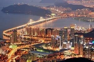 Aerial view of a city at night, showing an illuminated suspension bridge over water and numerous lit-up skyscrapers and roads.