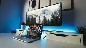 A modern desk setup with a silver laptop, a large monitor displaying a mountain landscape, and blue LED backlighting.