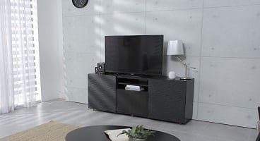 A modern living room with a large flat-screen TV on a dark, textured media console, a round coffee table with a plant, and sheer white curtains.