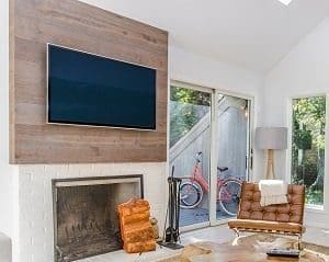 A living room with a TV mounted on a wooden wall above a white brick fireplace, a brown leather armchair, and a red bicycle visible through a glass sliding door.