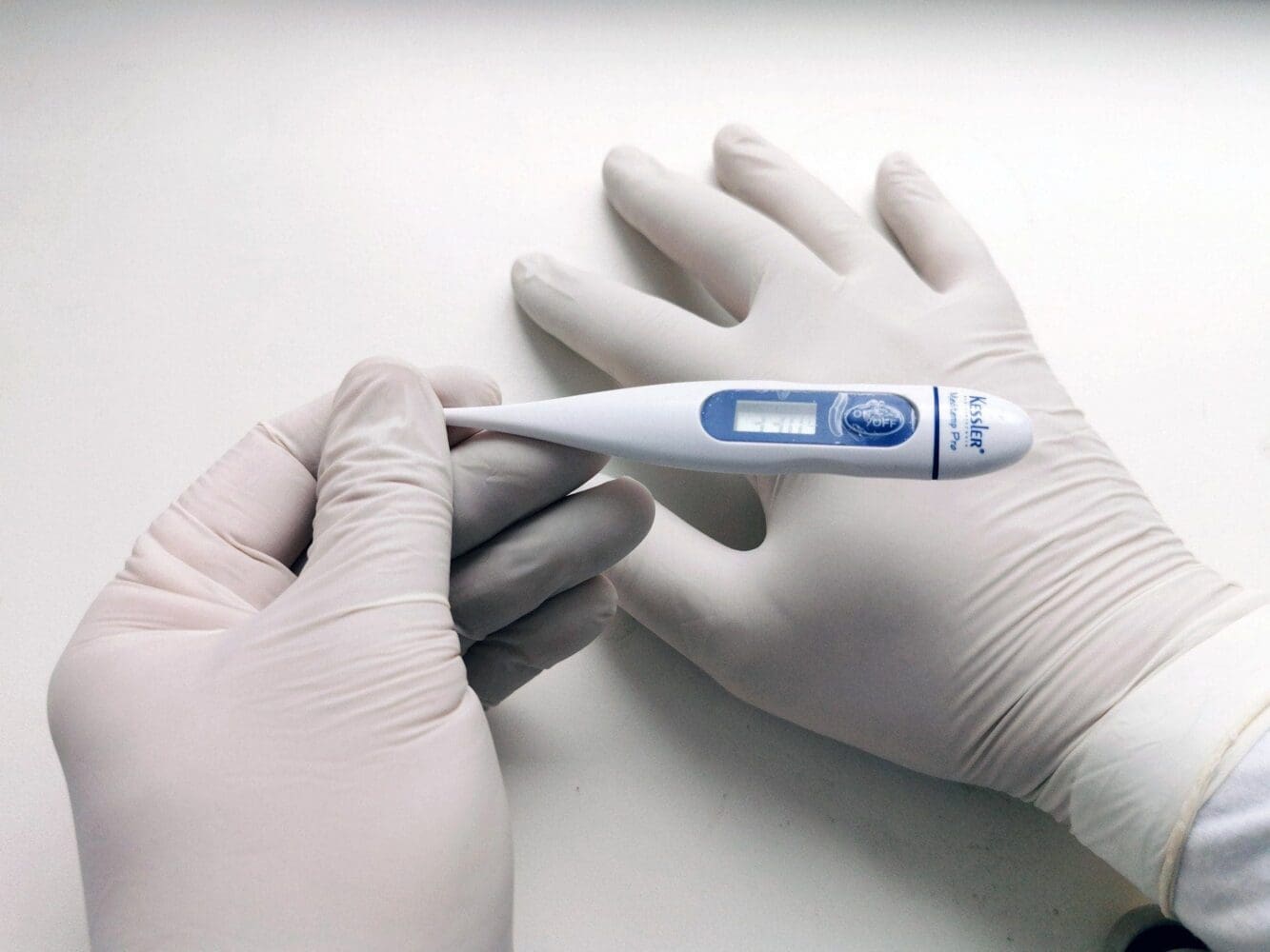 Two gloved hands hold a white and blue digital thermometer displaying '33.0' on its screen.