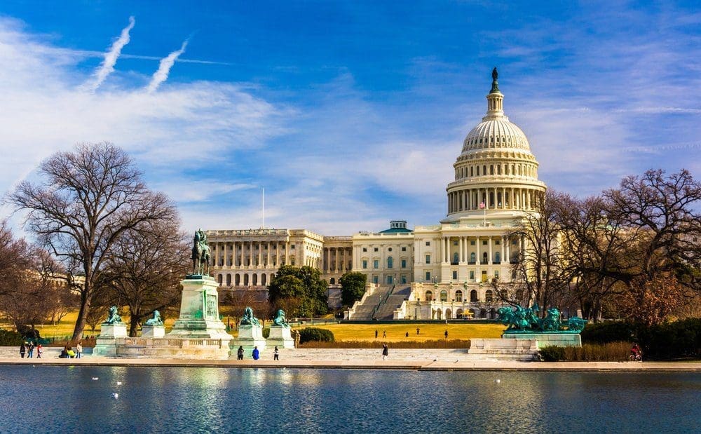 The U.S. Capitol Building with its dome, seen across a reflecting pool with statues and bare trees under a blue sky.