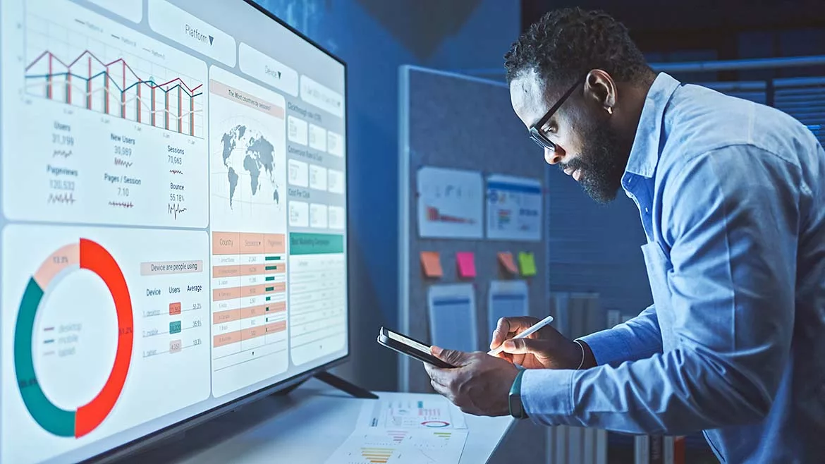 A man in glasses and a blue shirt analyzes data on a large monitor displaying charts, graphs, and metrics, while holding a tablet and stylus.