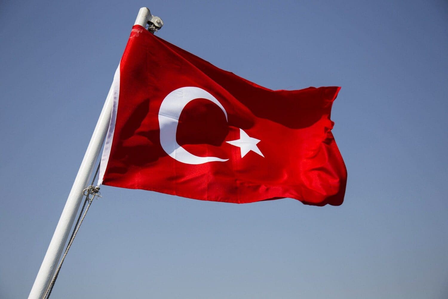 The red Turkish flag with a white crescent moon and star waves on a white flagpole against a clear blue sky.