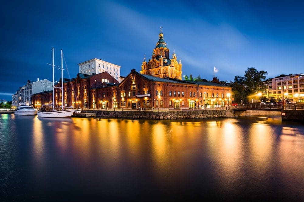 Night view of Helsinki's waterfront with illuminated brick buildings, docked boats, and the brightly lit Uspenski Cathedral reflecting in the calm water.