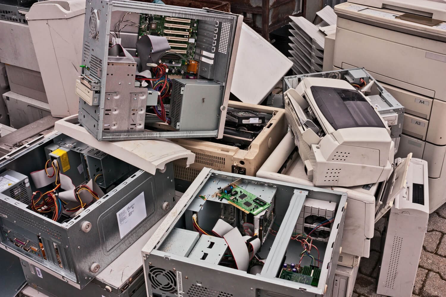 A large pile of old, beige and gray electronic waste, including computer towers with exposed components, printers, and scanners, stacked haphazardly.