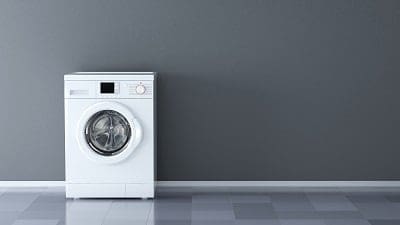 A white front-loading washing machine against a dark gray wall with a tiled floor.