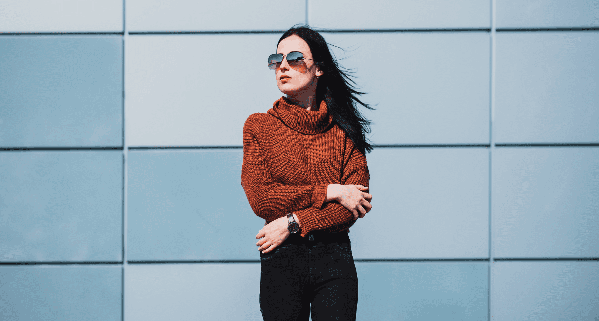 A woman in a rust-colored turtleneck and sunglasses stands in front of a blue and white paneled building.