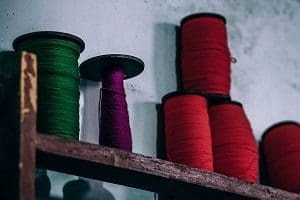 Green, purple, and red spools of thread are arranged on a wooden shelf against a light wall.