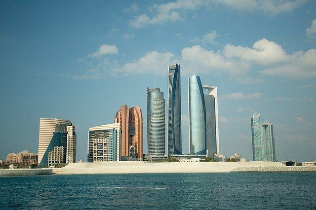 A panoramic view of a modern city skyline with numerous tall, contemporary skyscrapers along a waterfront, under a blue sky with white clouds.