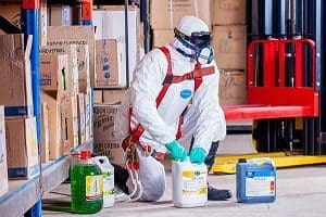 Person in a white hazmat suit, respirator, and gloves kneeling next to various chemical containers in a warehouse.