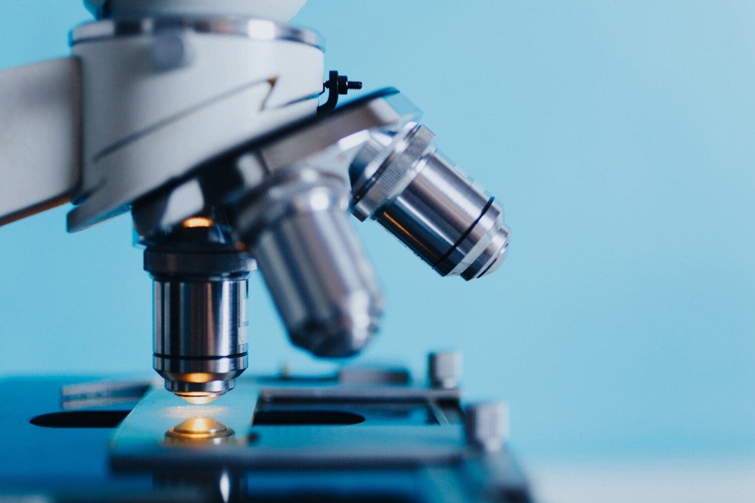 Close-up of a microscope with an objective lens focused on an illuminated sample on the stage, against a light blue background.