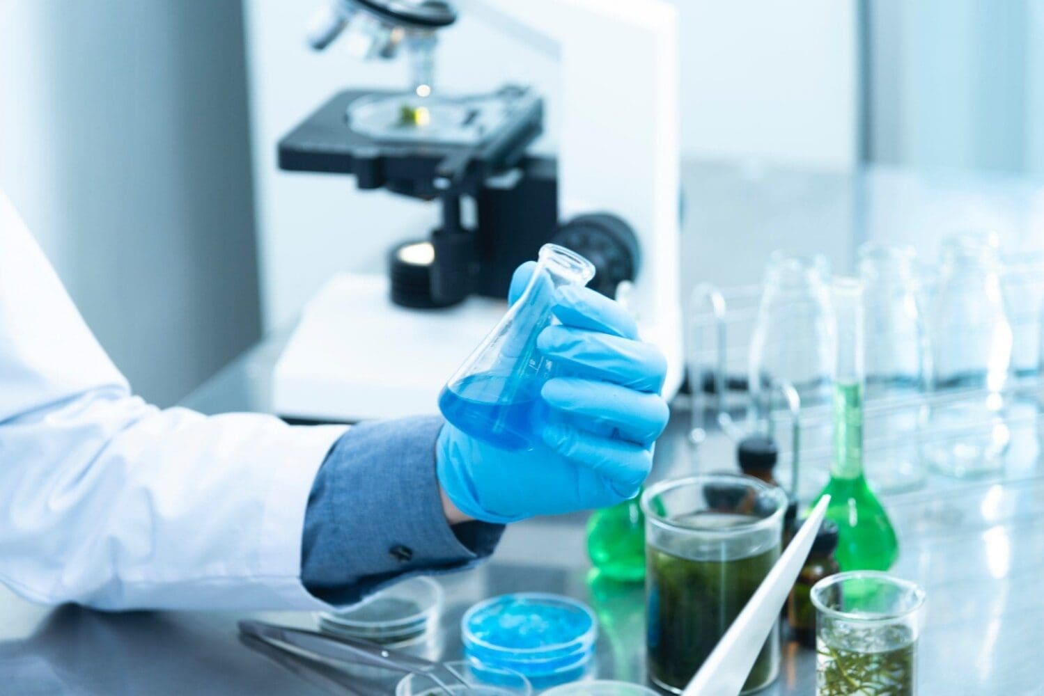 A gloved hand holds a flask with blue liquid in a laboratory, with a microscope and other beakers visible.
