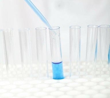 A pipette dispensing blue liquid into a test tube, surrounded by several empty test tubes in a white hexagonal rack.
