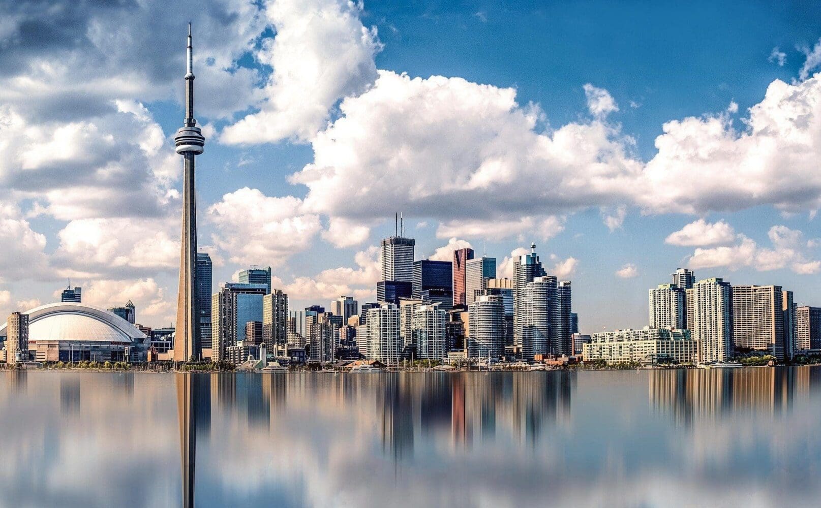 A panoramic view of the Toronto skyline, including the CN Tower and Rogers Centre, reflected in calm water under a blue sky with white clouds.