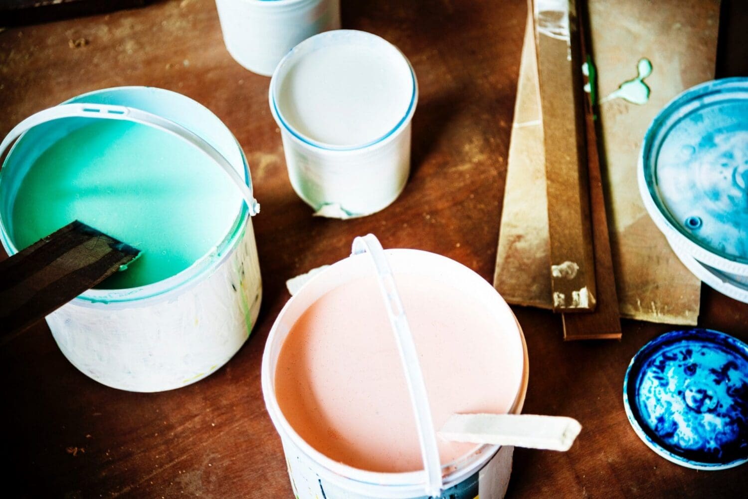 Overhead view of open paint cans with peach, turquoise, and white paint, stirring sticks, and paint-stained wooden pieces on a brown wooden surface.