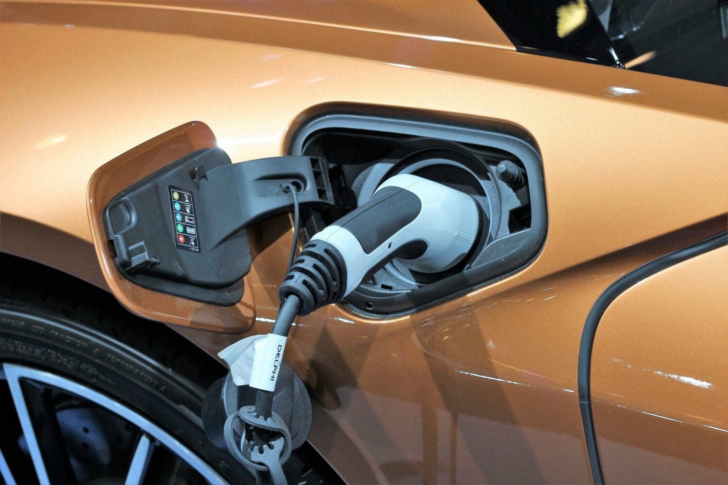 Close-up of a metallic orange-bronze electric car with a white and black charging plug inserted into its open charging port. A 'DELPHI' tag is visible on the cable.