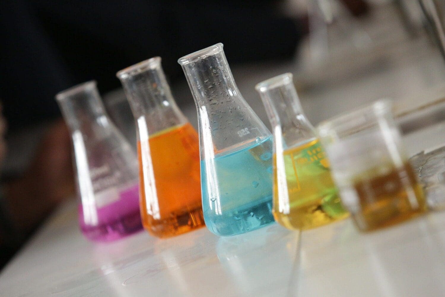 Five laboratory flasks and beakers containing purple, orange, blue, yellow, and brownish-yellow liquids, arranged diagonally on a light surface.