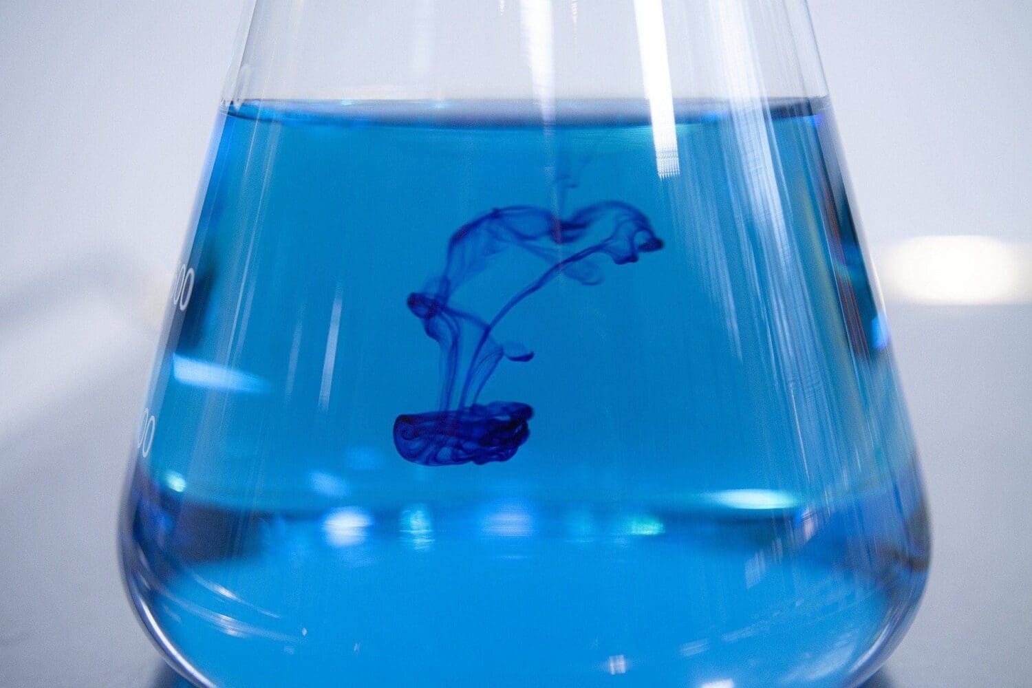 A clear glass Erlenmeyer flask filled with bright blue liquid, with darker blue dye diffusing and swirling downwards into it.