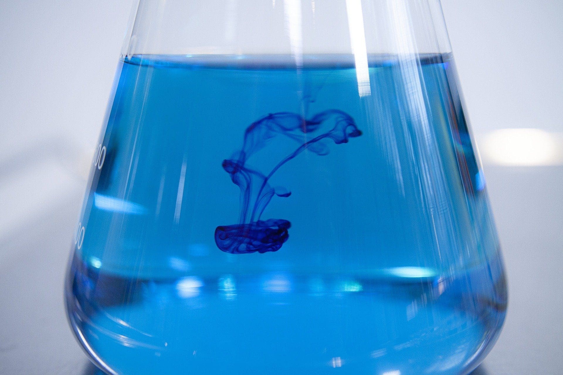 A clear glass Erlenmeyer flask filled with bright blue liquid, with darker blue dye diffusing and swirling downwards into it.