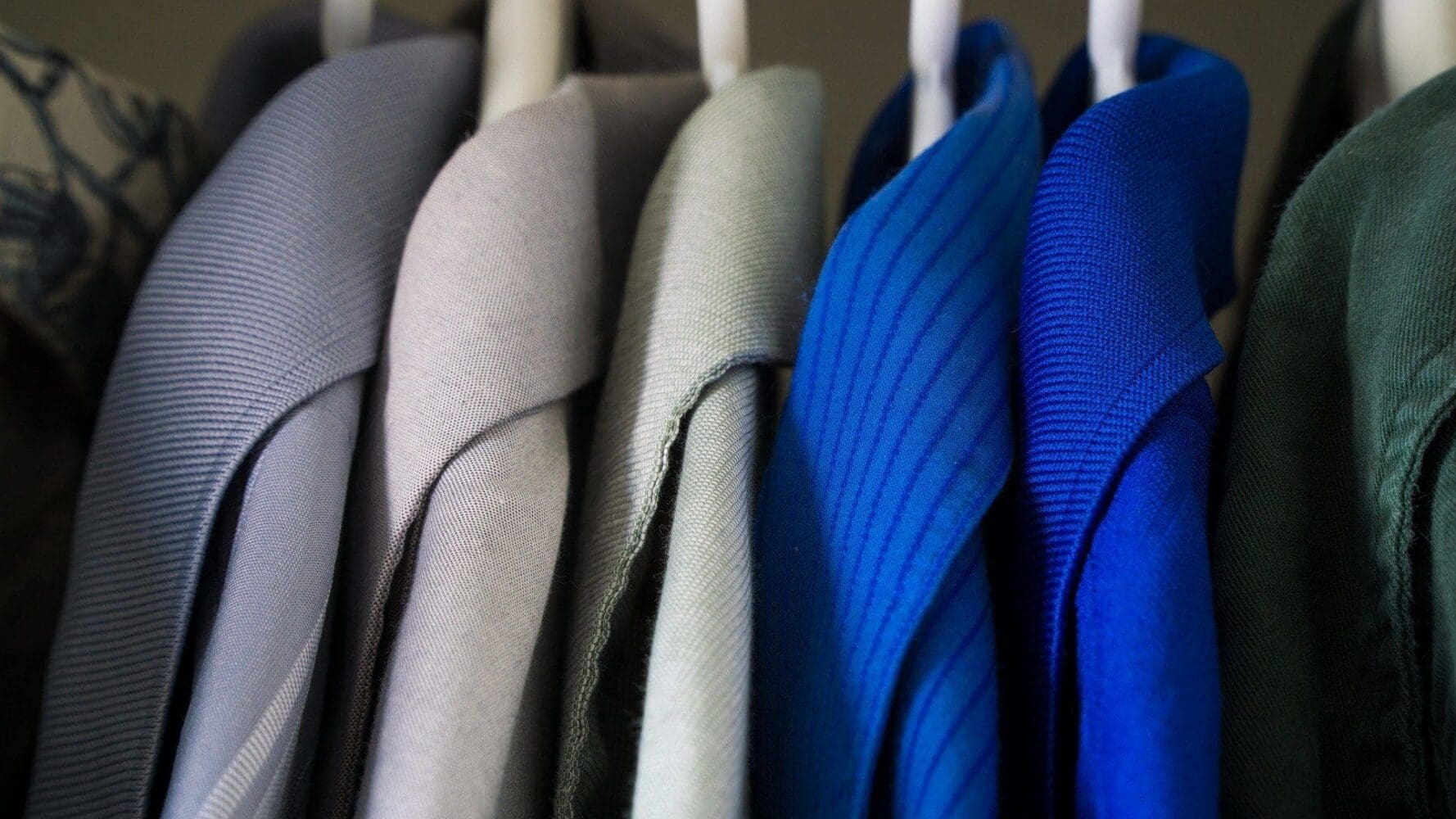 Close-up of several shirts in various shades of blue, grey, green, and light blue, hanging neatly on white hangers in a closet.