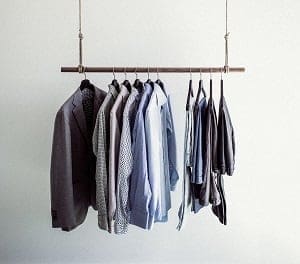 Men's clothing, including a dark gray blazer and several button-down shirts in blue, gray, and white, hanging on a suspended metal rack against a plain white wall.