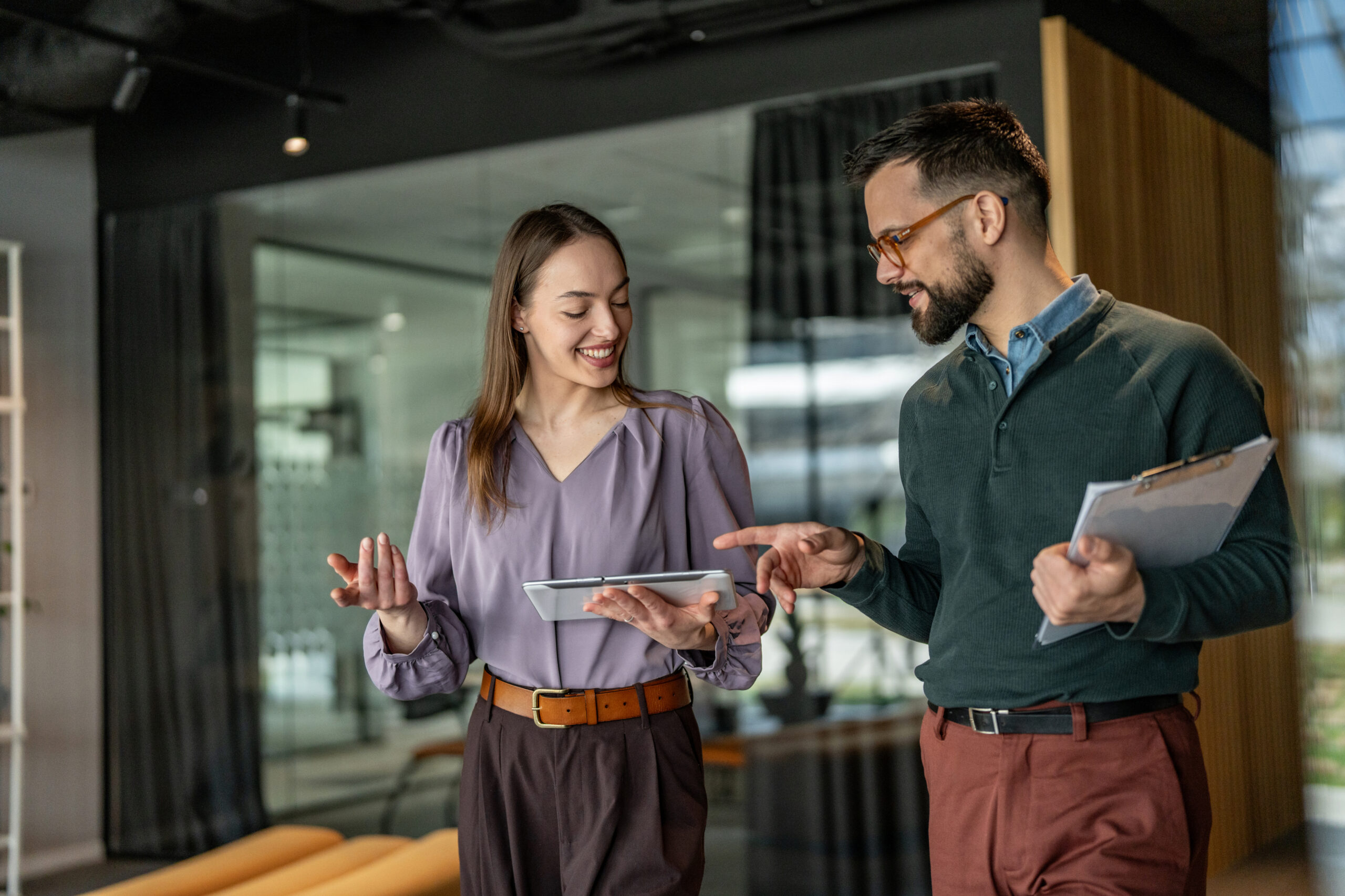 A smiling woman and man in an office, looking at a tablet. The man points at the screen while the woman holds it.