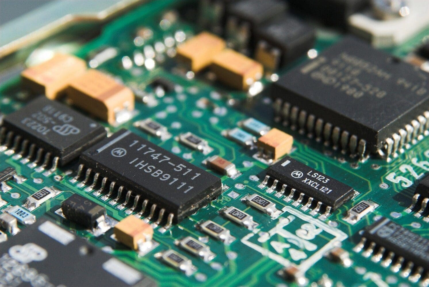 Close-up of a green circuit board with various black integrated circuit chips, orange capacitors, and small resistors. Text on chips reads '11747-511 IHSB9111' and 'LS123 XXCL 121'.
