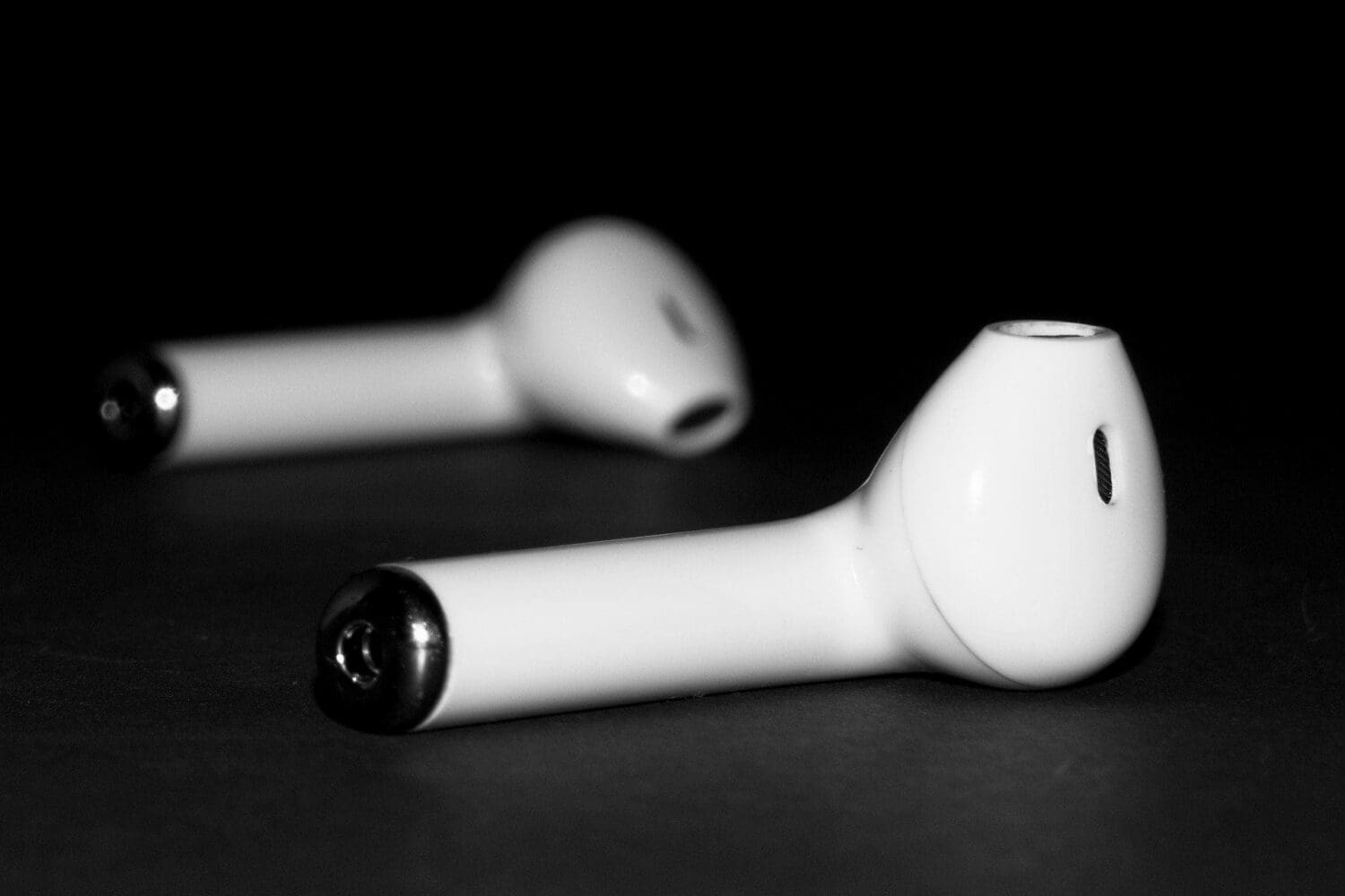 Two white wireless earbuds, one in focus in the foreground, rest on a dark surface in a black and white image.