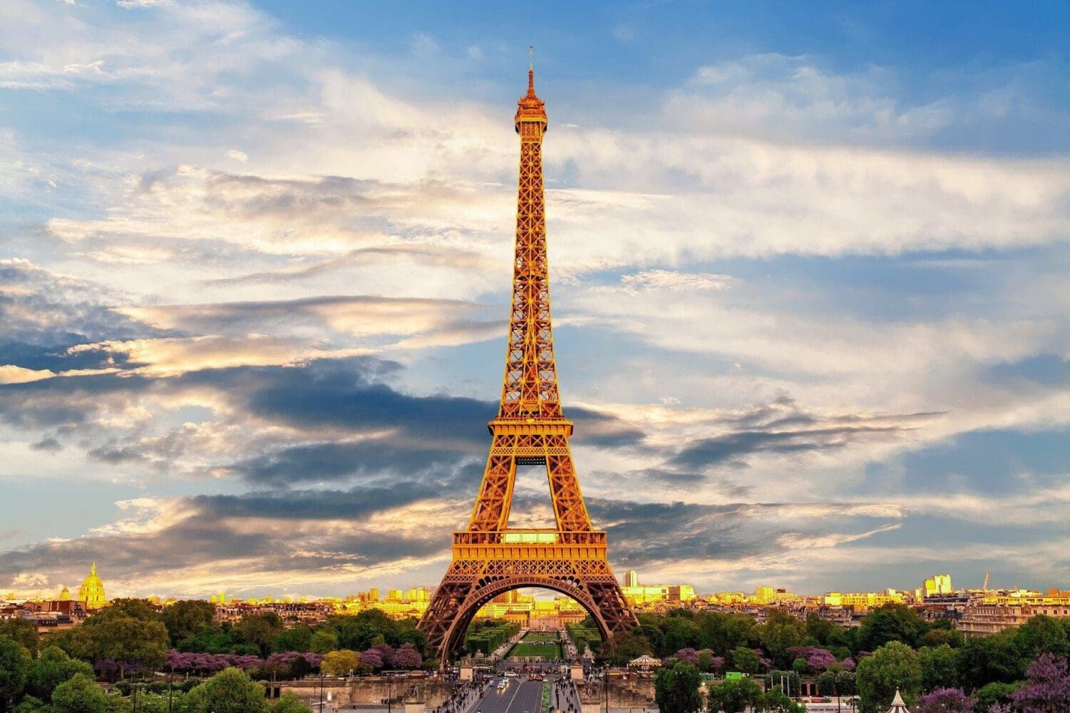 The Eiffel Tower stands tall and golden against a dramatic, cloudy sunset sky, with the city of Paris and lush trees in the foreground.