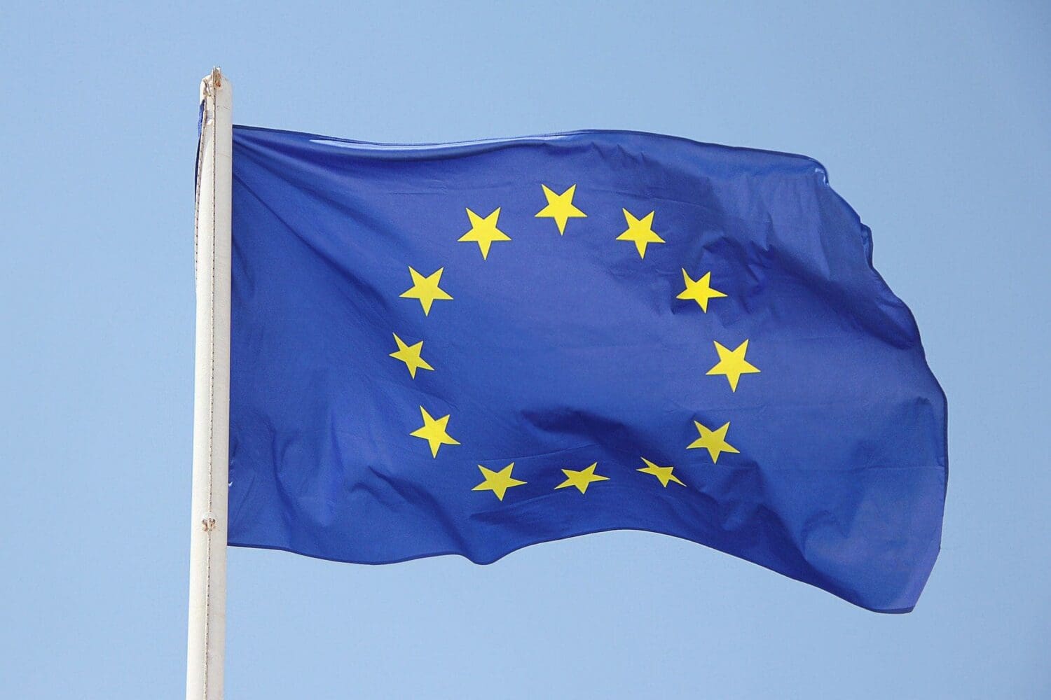 The European Union flag, blue with 12 yellow stars, waving on a white flagpole against a clear blue sky.