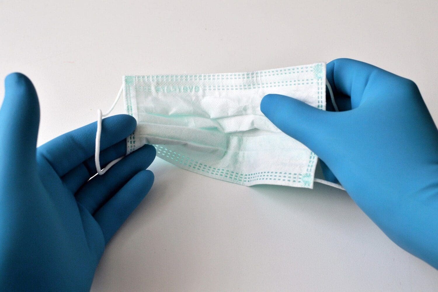 Two hands in blue medical gloves holding a light blue disposable surgical mask.