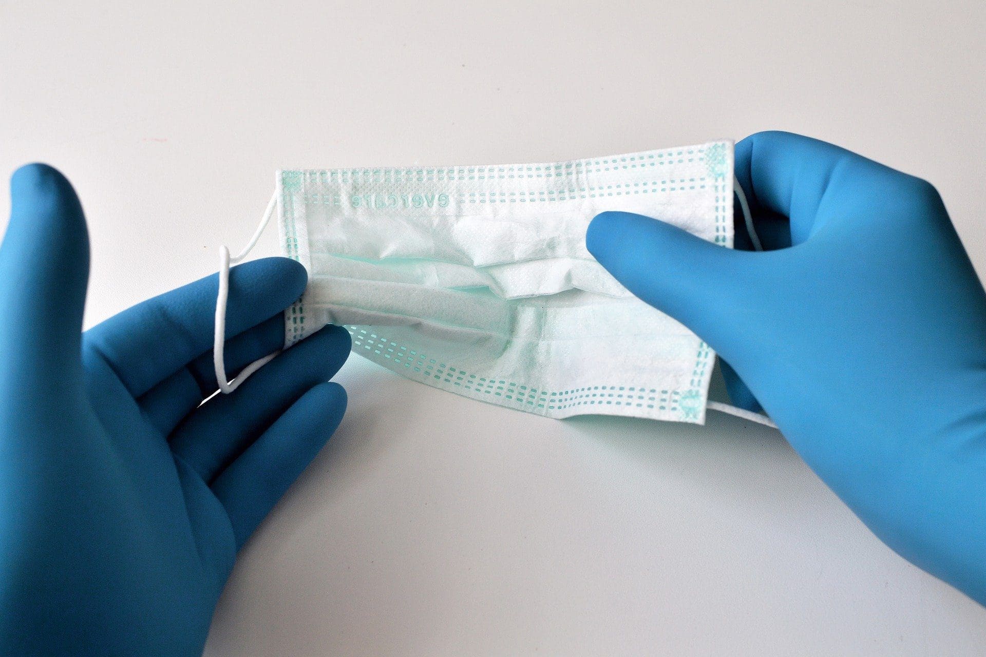 Two hands in blue medical gloves holding a light blue disposable surgical mask.